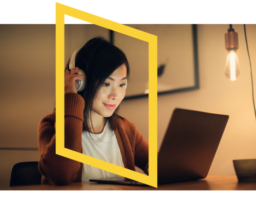 A woman wearing headphones sits at a desk using a laptop, framed by a yellow geometric shape. A hanging lightbulb glows in the background, creating a warm, focused atmosphere.