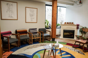 A cozy waiting room with four chairs, a rocking chair, a small round glass table, and a decorative rug. There’s a fireplace, potted plants by the window, and framed art on the walls. Natural light enters through the window.