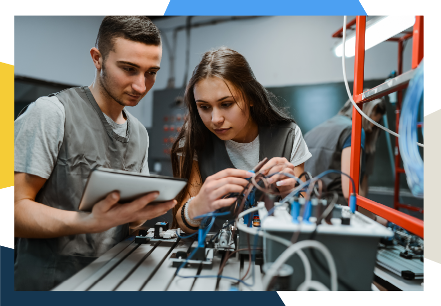 Two young people in gray vests work together on electronics at a lab table. One holds a tablet, while the other adjusts wires connected to a circuit board, with various equipment visible around them.